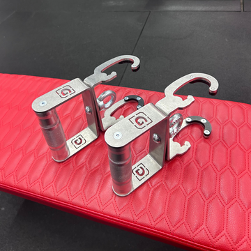 A pair of stainless steel G-shaped handles with knurling, placed on a red padded surface, presumably a gym bench.