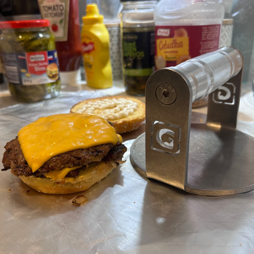 Hamburger with cheese on a bun next to a burger press on a kitchen counter with condiments in the background.