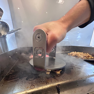 Hand using a Gympin Steel Burger Patty Presson a hot surface with food residue