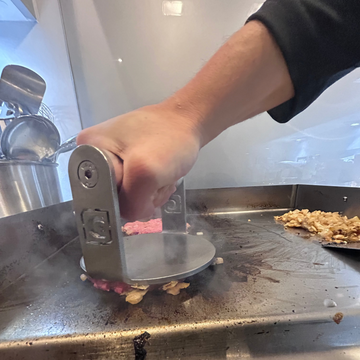 Person using a burger patty press on ground meat on a metal surface.